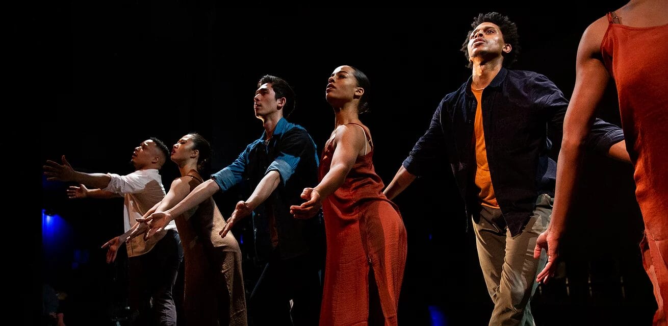 Dancers on stage performing a contemporary routine with expressive gestures and coordinated formation.